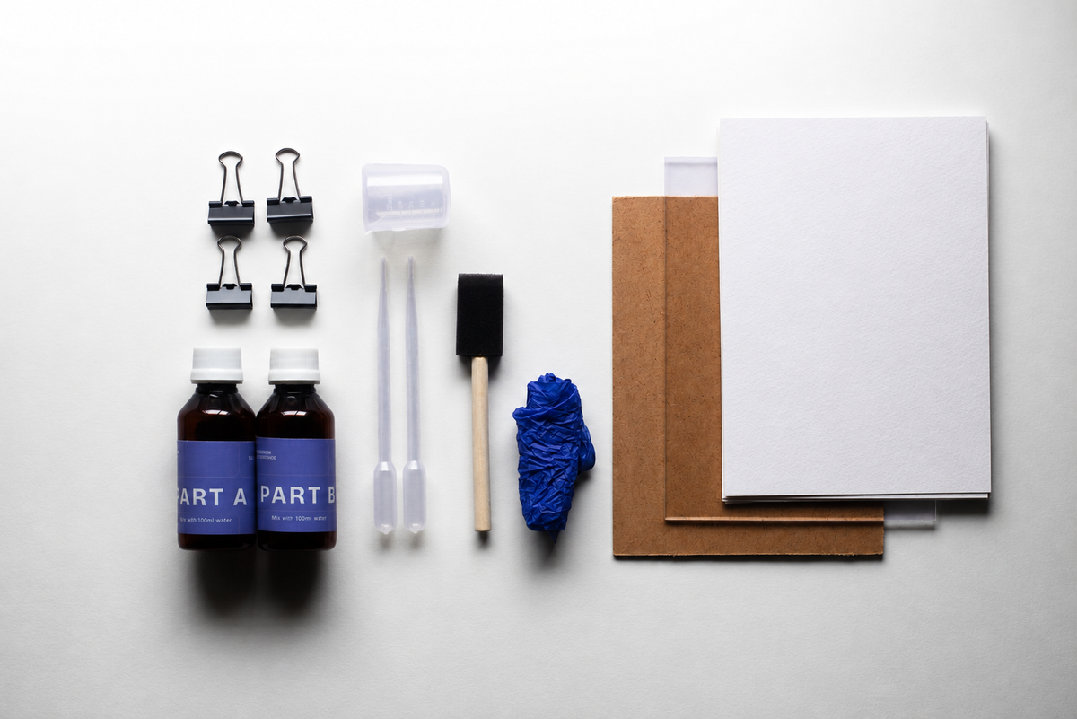 A flat-lay of a DIY craft or repair kit on a white background, including two small bottles labeled PART A and PART B, four binder clips, two plastic syringes, a foam brush, a blue cloth, brown and white paper sheets, and a white card.