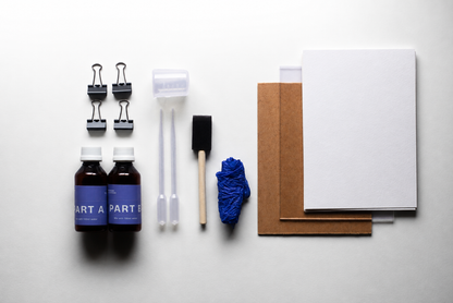 A flat-lay of a DIY craft or repair kit on a white background, including two small bottles labeled PART A and PART B, four binder clips, two plastic syringes, a foam brush, a blue cloth, brown and white paper sheets, and a white card.